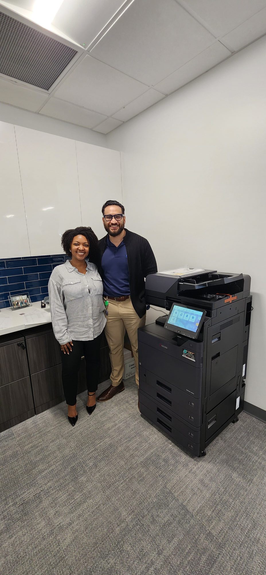 A smiling professional team from a Broward commercial realty management company standing next to their upgraded Kyocera TASKalfa office hub following a seamless installation by STAT Business Systems.