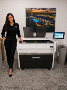 KIP Wide Format Office Printer From STAT - STAT Business Systems - STAT Business Systems Woman in black business attire standing beside a large white and black multifunction printer in an office.