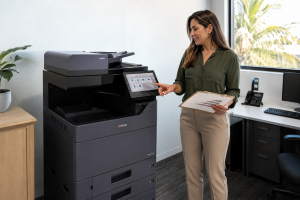 Calculating Monthly Print Volume Before Leasing a Copier - STAT Business Systems - STAT Business Systems Woman in a green blouse uses a Kyocera multifunction printer, touching the touchscreen while holding a clipboard with charts in an office.